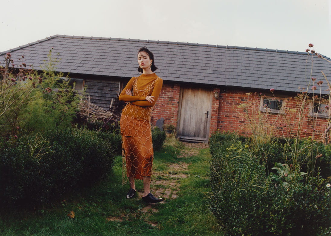 A model stands in front of a farmhouse in a chain mail dress