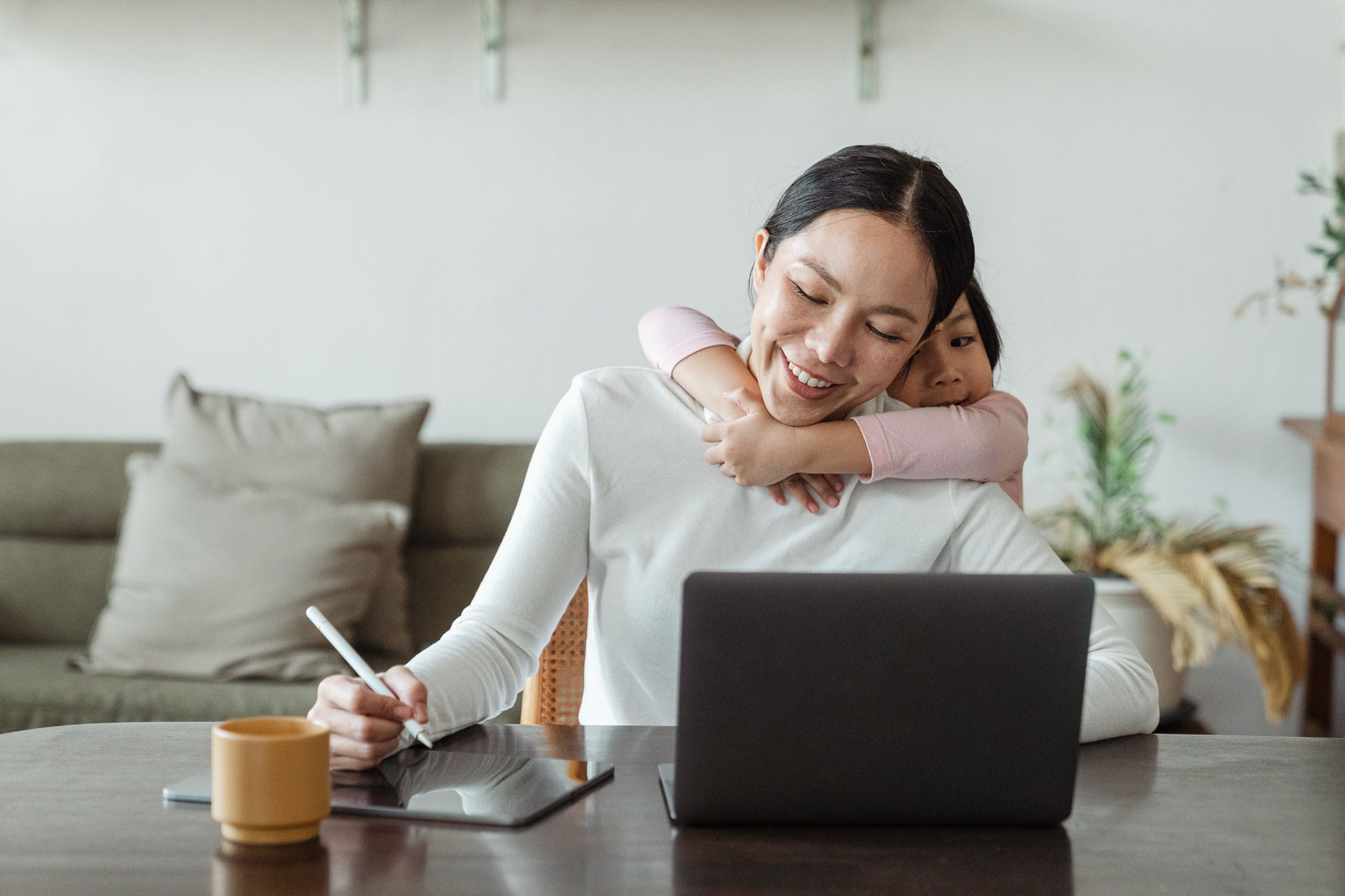 A mother working on a laptop with a child hugging her in celebration of Mother's Day